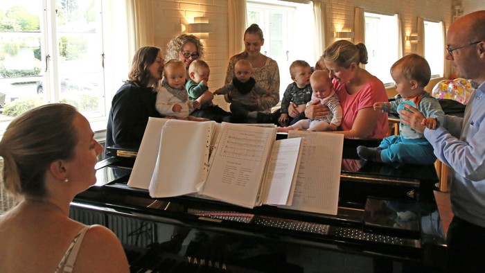 En gruppe af voksne og børn synger sammen ved et piano i et lys og rummeligt rum