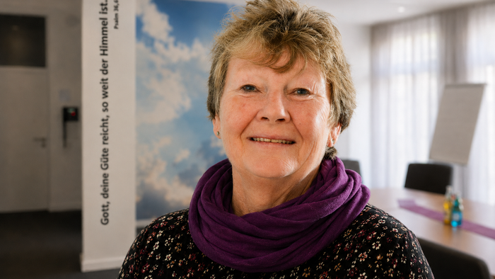 Porträt einer freundlich lächelnden Frau mit kurzem Haar und violettem Schal in einem hellen Gemeinderaum. Im Hintergrund sind ein Wandzitat mit Bibelvers, ein Himmelmotiv sowie ein Besprechungstisch zu sehen. Die Aufnahme wirkt warm, ruhig und einladend.