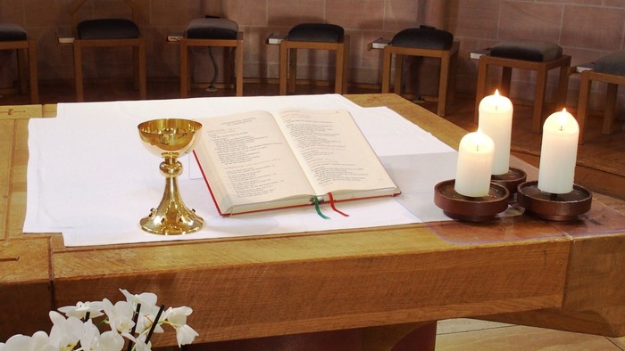 Religiöser Altarschmuck mit einem aufgeschlagenen Buch, Kelch, Kerzen und Blumengesteck auf einem Holztisch.