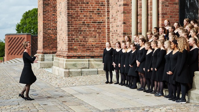 En gruppe korister står i en linje og synger med en dirigent foran en mur af røde mursten.