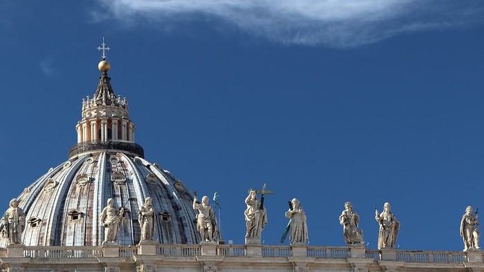 Das Bild zeigt den Kuppelbau des Petersdoms mit zahlreichen Statuen vor blauem Himmel.