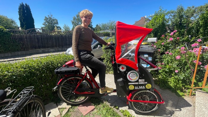 Eine Person fährt ein rotes Lastenrad mit seitlichem Anbau, das in der Nähe von üppigem Grün und blühenden Pflanzen geparkt ist.