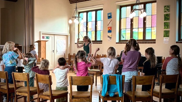 Die Kinder versammeln sich aufmerksam in einem klassenzimmerähnlichen Raum und hören einem Lehrer an einem hölzernen Pult zu.
