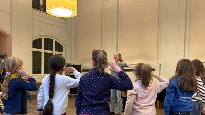 Kinder in einem Klassenzimmer oder einer Kapelle, die die Hände heben, während ein Erwachsener steht und sie zu unterrichten oder mit ihnen zu interagieren scheint.