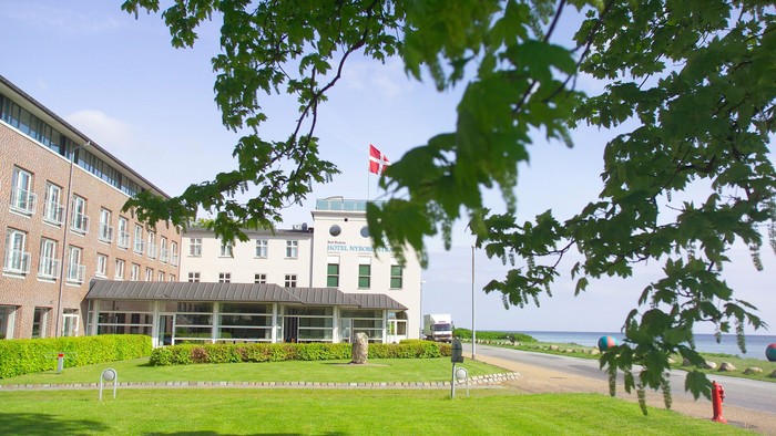 Et stort hvidt hotel ved stranden med dansk flag på en stang.