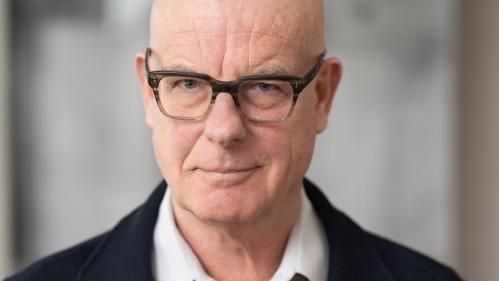 Ein glatzköpfiger Mann mit Brille, der einen dunklen Blazer über einem weißen Hemd trägt, posiert formell.