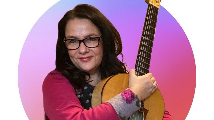 Eine Person mit Brille spielt eine klassische Gitarre mit einer gemütlichen gestrickten Hülle.