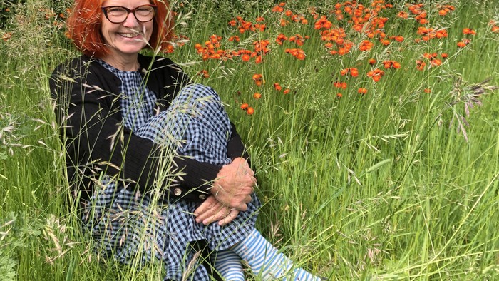 En person med lyserødt hår sidder smilende i en mark med højt græs og levende orange blomster.