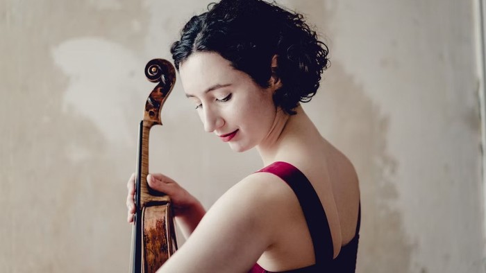 Eine Geigerin mit lockigem Haar hält ein Cello, elegant gekleidet in ein dunkles Kleid, versunken in musikalische Kontemplation.