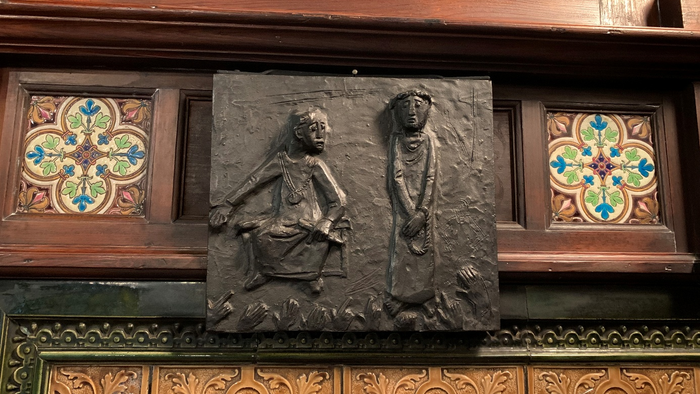 Reliefskulptur, die zwei skelettierte Figuren in einer Holztafel mit dekorativen floralen Rändern darstellt.