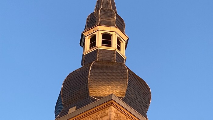 Ein historischer Steinkirchturm mit zwei Uhren und einem spitzen Turmhelm unter einem klaren blauen Himmel.