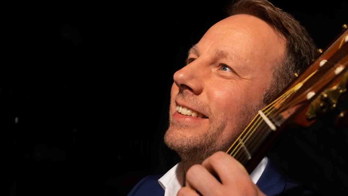 Ein lächelnder Mann spielt eine akustische Gitarre in einer dunklen Umgebung, gekleidet in einen Anzug und ein weißes Hemd.