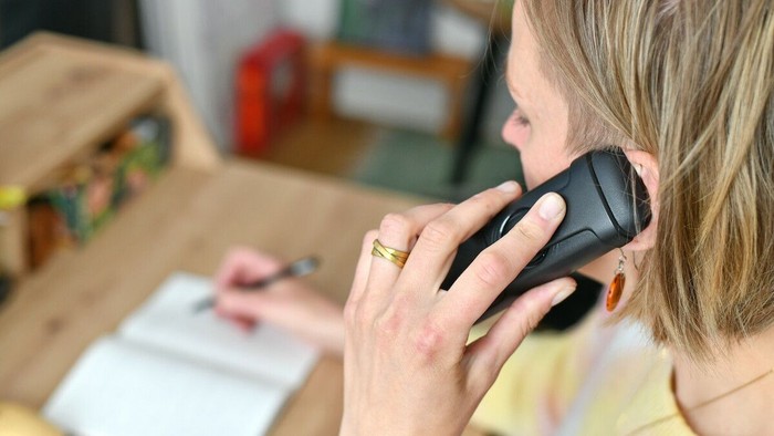 Eine Person spricht an einem schnurlosen Telefon, während sie Notizen auf ein Blatt Papier an einem Holztisch schreibt.