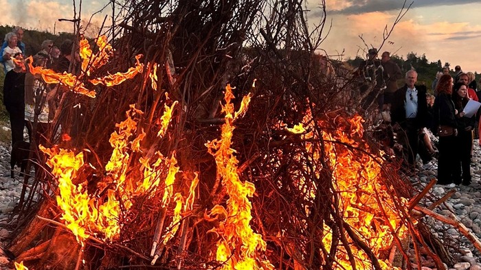 En gruppe mennesker står omkring en stor bål på en stenstrand ved solnedgang