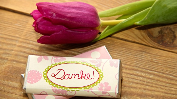 Eine rosa Tulpe und verpackte Pralinen mit dem Aufkleber „Danke!“ auf einer Holzoberfläche.