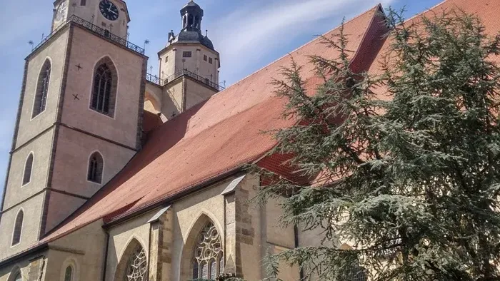 Historische Kirche mit hohem Turm und rotem Ziegeldach, eingerahmt von einem immergrünen Baum und angrenzenden Gebäuden.
