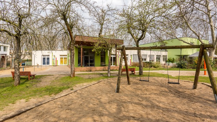 Im Bild ist ein Spielplatz mit Schaukeln und einer grünen Überdachung zu sehen, umgeben von Bäumen und einem Gebäude im Hintergrund.