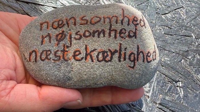 En hånd holder en glat sten med håndskrevne danske ord om tilfredshed og venlighed.