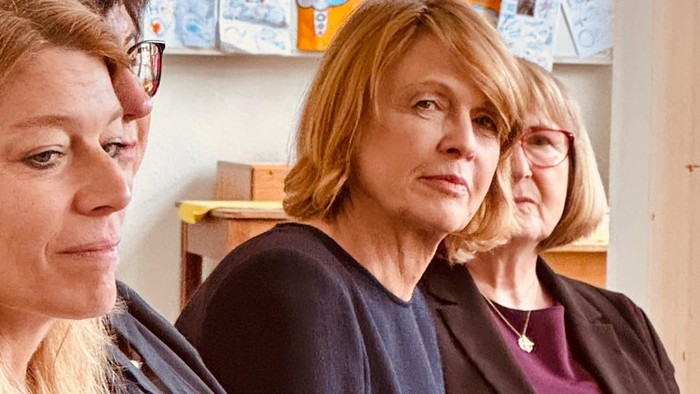 Drei Frauen sitzen aufmerksam in einem Klassenzimmer oder Besprechungsraum mit bunten Bildungsplakaten im Hintergrund.