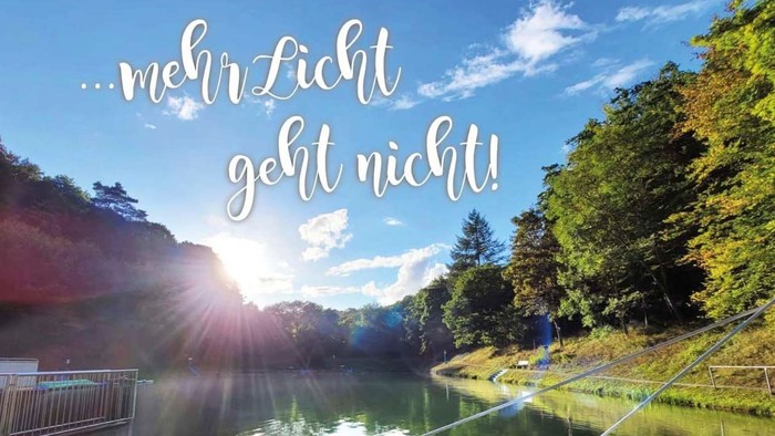 Sonnenbeschienene Seeszene mit üppigem Grün und dem deutschen Spruch *„Mehr Licht geht nicht!“* darübergelegt.