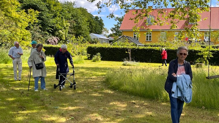 Gruppe af mennesker, der går og nyder et naturskønt udendørs haveområde med et farverigt hus i baggrunden.