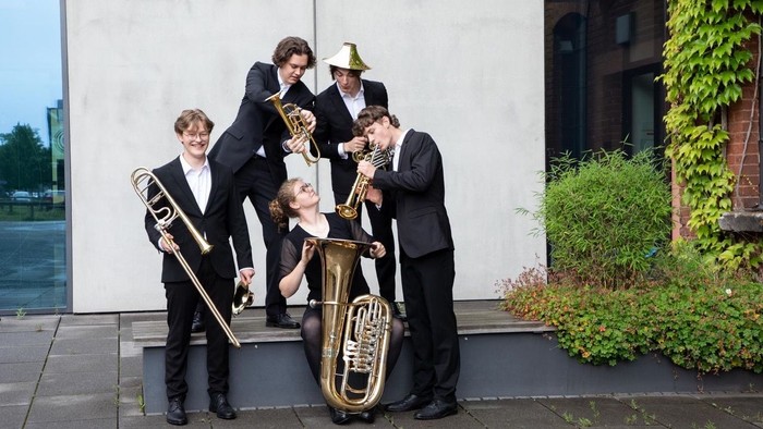 Eine Gruppe von Musiker:innen in formeller Kleidung, die im Freien mit Blechblasinstrumenten posieren.