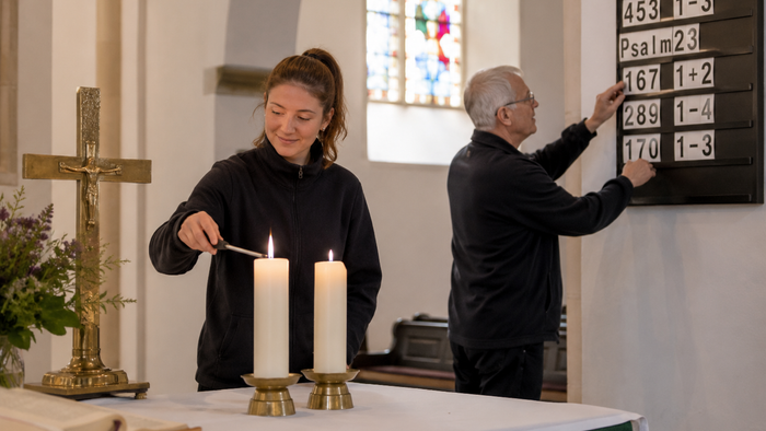 Eine junge Küsterin zündet am Altar einer Kirche Kerzen an, während im Hintergrund ein älterer Küster die Liednummern an einer traditionellen Liedtafel austauscht. Auf dem Altar stehen Kerzen, ein Kreuz und ein aufgeschlagenes Buch. Tageslicht fällt durch