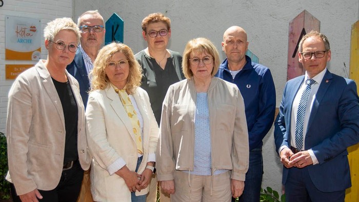 Gruppe von sieben Personen, die draußen in der Nähe von farbenfroher architektonischer Beschilderung und einem Gebäude posieren.