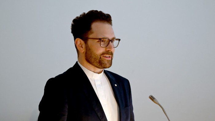 Ein Mann in einem Anzug steht an einem Mikrofon und hält eine Rede.