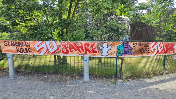 Bunter Banner zur Feier von „St. Mauritan Kolbe 50 Jahre“ mit festlicher Graffiti auf einem Gehweg in der Nähe eines Zauns.