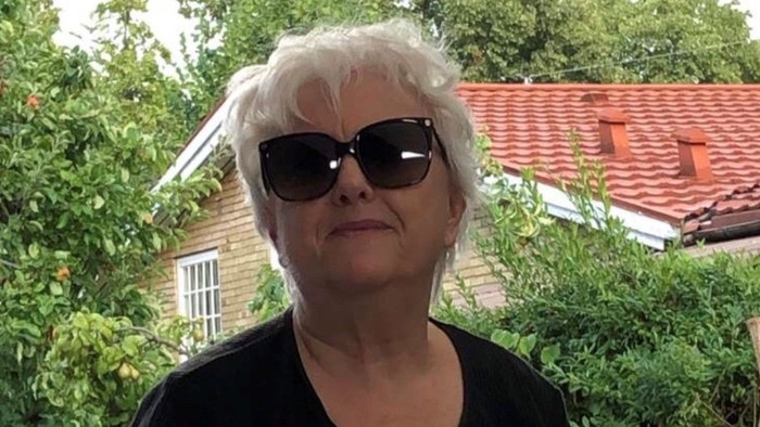 En person med hvidt hår iført solbriller står udendørs nær frodig grønne omgivelser og et hus.