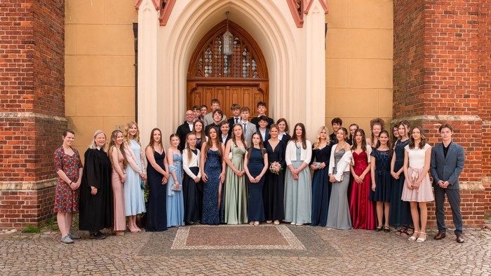 Ein formelles Gruppenfoto von Absolventen in eleganter Kleidung, die vor dem Eingang eines historischen Backsteingebäudes stehen.
