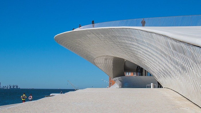 Ein modernes, geschwungenes weißes Gebäude mit begehbarem Dach. Im Hintergrund Meer.