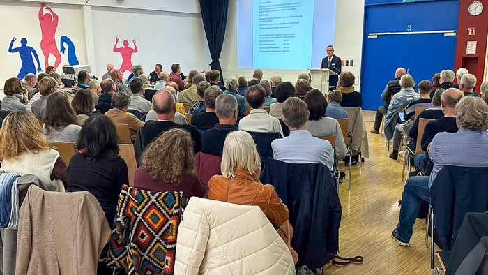 Ein Publikum sitzt in Reihen und hört einem Redner an der Vorderseite zu.