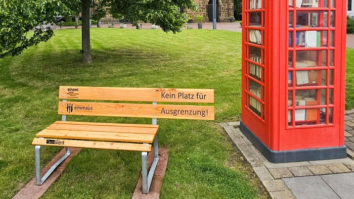 Eine Bank mit einem verkürzten Sitzteil steht neben einer roten Telefonzelle vor einer Kirche