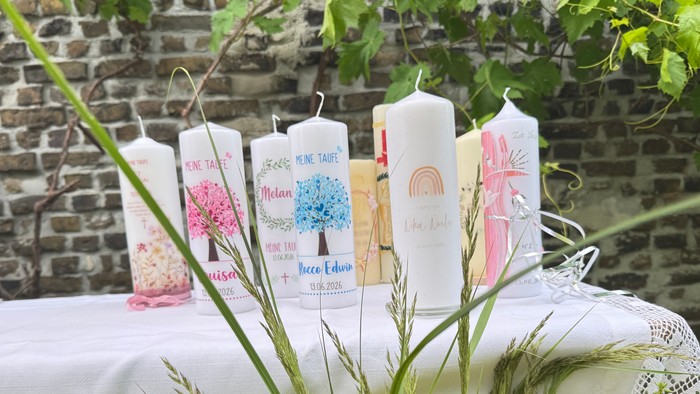 Verzierte Kerzen und Blumen, arrangiert auf einem rustikalen Tischtuch neben einer Backsteinmauer.