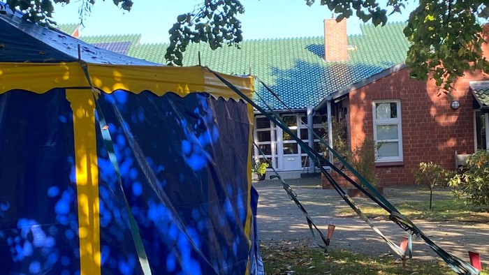 Ein blau-gelbes gemustertes Zelt, das im Freien in der Nähe eines Backsteingebäudes aufgebaut ist, mit Herbstblättern auf dem Boden.