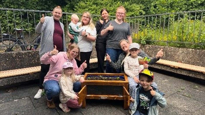 Eine fröhliche Familiengruppe posiert im Freien neben einem hölzernen Bienenstock und zeigt Daumen-hoch-Zeichen.