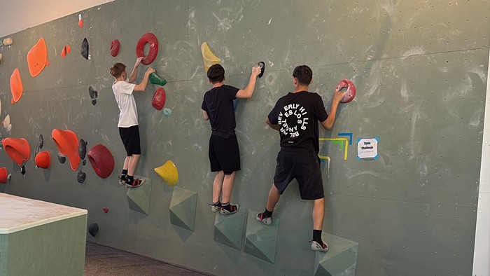 Zwei Personen, die an einer Indoor-Kletterwand mit verschiedenen farbigen Griffen und Tritten klettern.