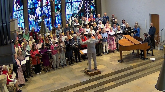 Ein Chor tritt in einer Kirche mit bunten Glasfenstern auf, geleitet von einem Dirigenten und einem Pianisten.