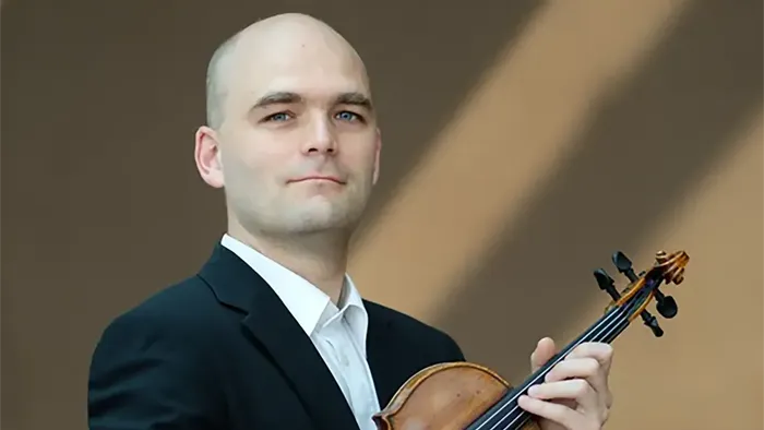En skaldet mand i jakkesæt holder og spiller på en violin mod en neutral baggrund.