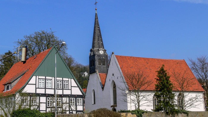 Eine weiße Doppelkirche mit einem dunklen Turm, davor ein Fachwerkhaus, aus der Entfernung über eine Mauer gesehen