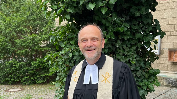 Ein lächelnder Mann in geistlichem Gewand mit einem Kreuzemblem steht im Freien neben Grünpflanzen.