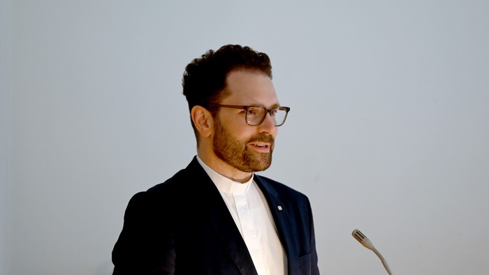 Ein Mann in einem Anzug steht an einem Mikrofon und hält eine Rede.