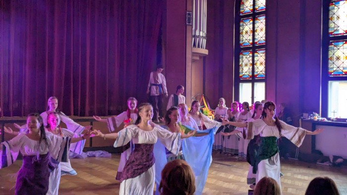 Eine Gruppe von Tänzern in historischer Kleidung tritt in einem prächtigen Saal mit bunten Glasfenstern auf.