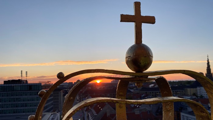 Udsmykket gylden krone med globus og kors sat op mod en bylandskab ved solnedgang.
