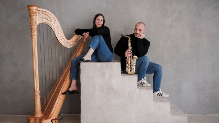 Zwei Musiker posieren mit einer Harfe und einem Saxophon auf einer minimalistischen Treppenkulisse.