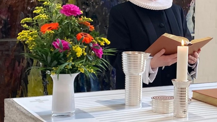 En person i mørkt tøj holder en bog og står ved et bord med blomster og lysstake.