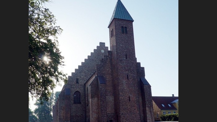 Historisk murstenskirke med højt spir i et fredeligt, grønt landskab.