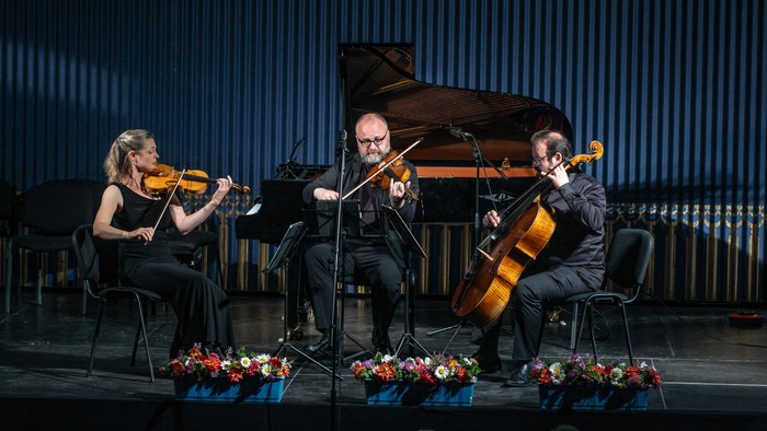 Musikere, der optræder i et kammermusikensemble med violin, cello og klaver på en scene pyntet med blomsterarrangementer.